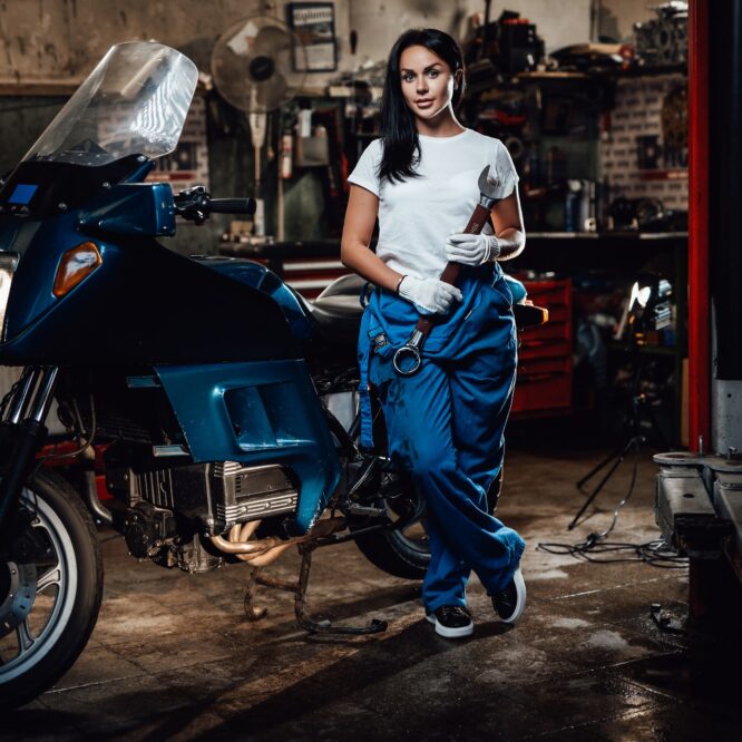 Beautiful brunette woman in blue overalls posing next to a sportbike in garage or workshop