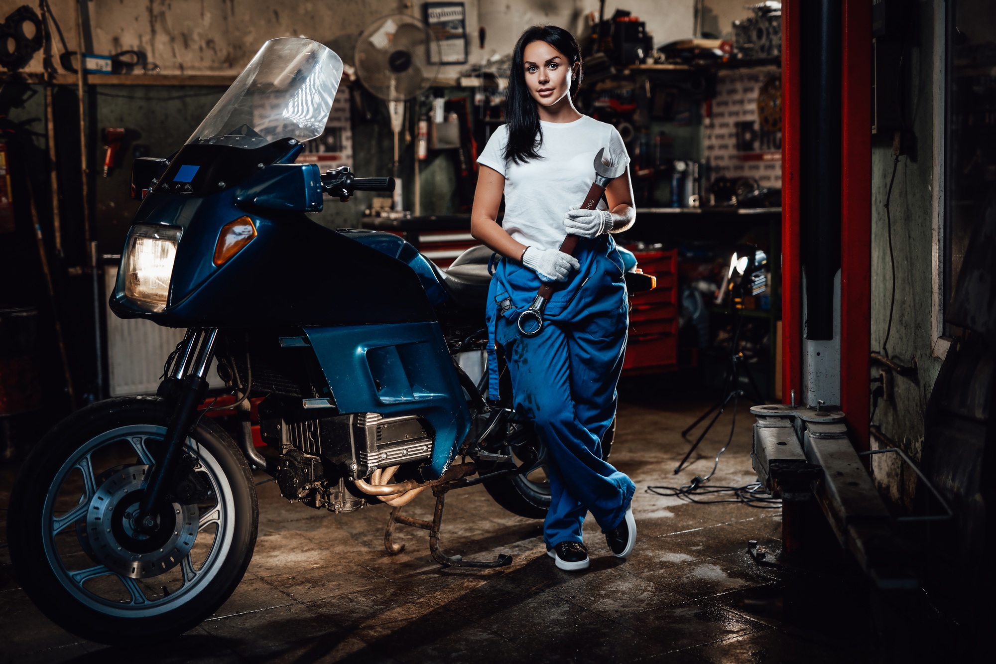 Beautiful brunette woman in blue overalls posing next to a sportbike in garage or workshop